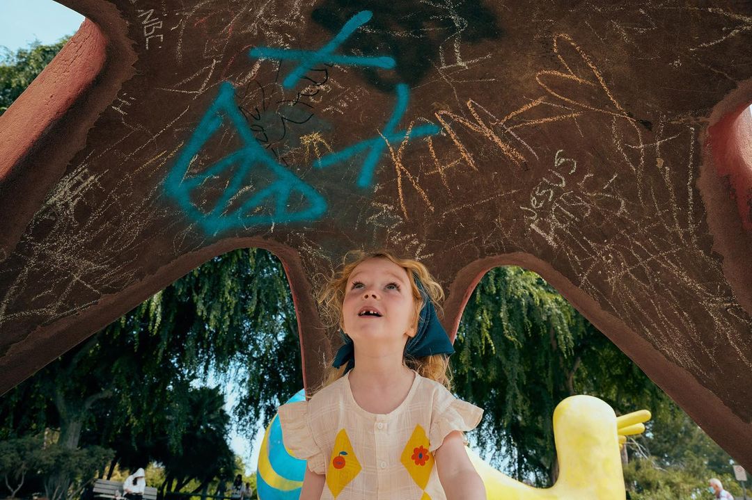 Low-angle view of a red-haired child gazing upwards at graffiti on a park structure with trees and people in the background on a sunny day.