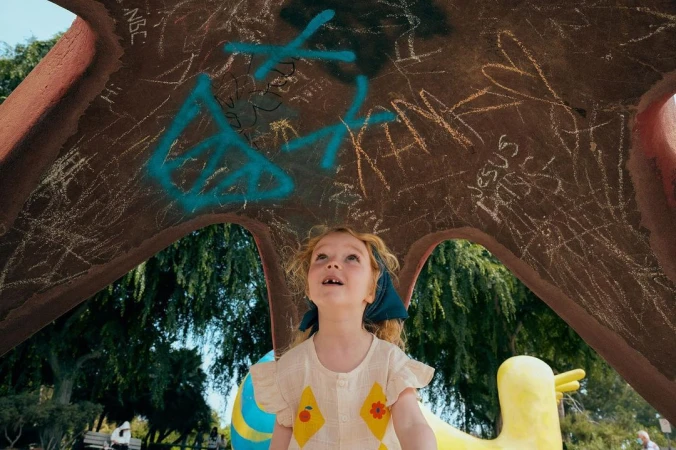 Low-angle view of a red-haired child gazing upwards at graffiti on a park structure with trees and people in the background on a sunny day.