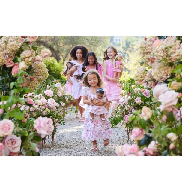 A group of young girls in pink dresses walk through a flower garden, each holding a doll, enjoying a day out surrounded by roses.