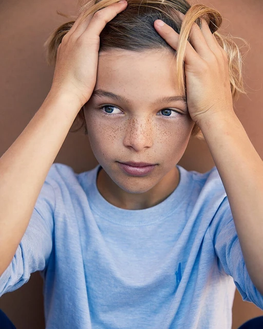 Portrait of a young boy with blond hair and freckles, holding his hands near his head, captured in soft, natural light.