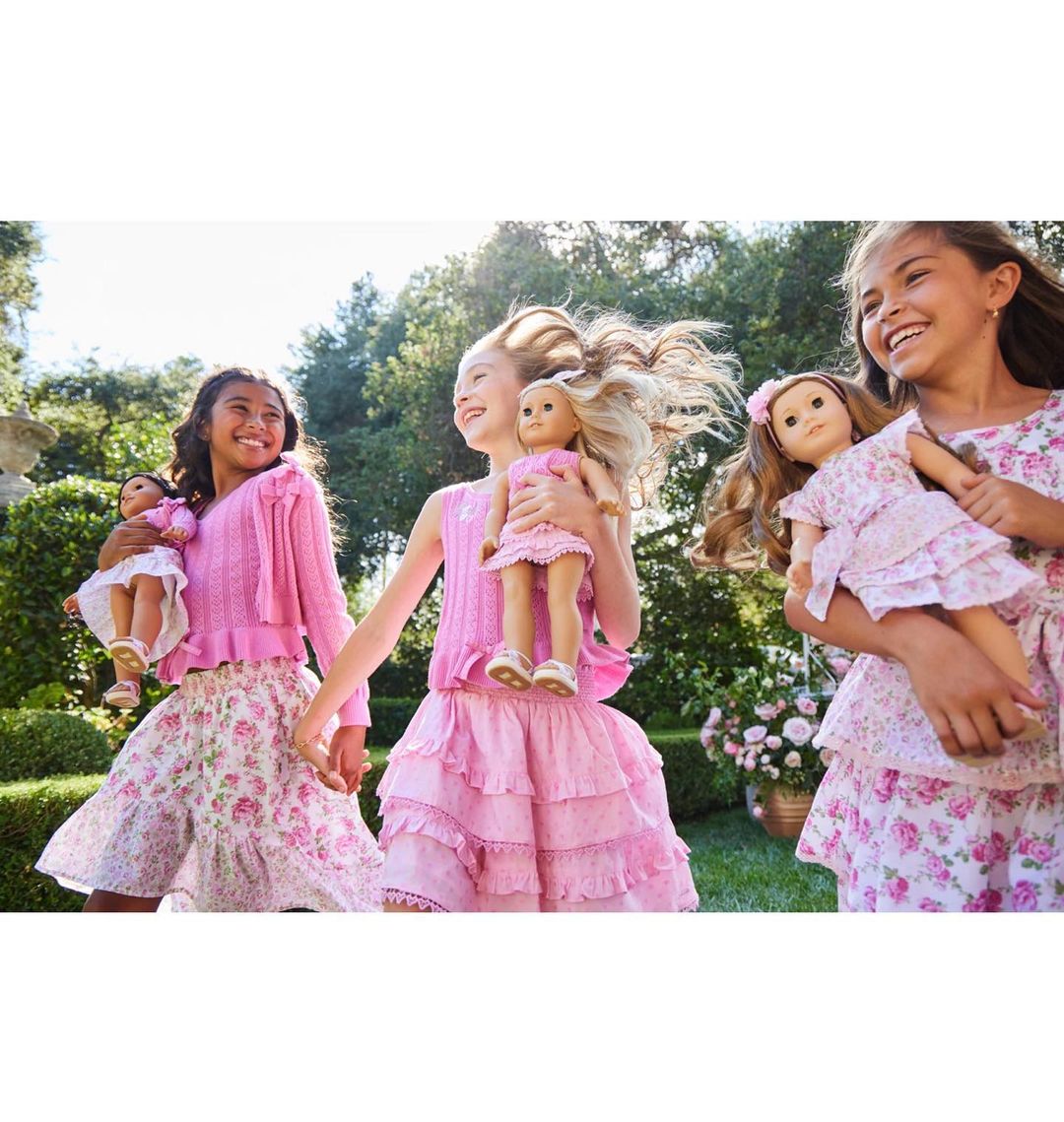 Three young girls in pink floral dresses hold matching dolls in a sunlit outdoor setting, smiling and holding hands.