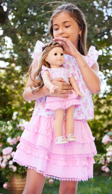 A young girl in a floral dress outdoors holds a matching doll, both in pink and white floral outfits, against a leafy green background.