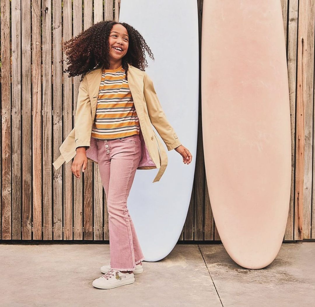 Smiling young girl with curly hair poses in front of surfboards wearing a tan jacket and pink pants, outdoor setting, full shot.