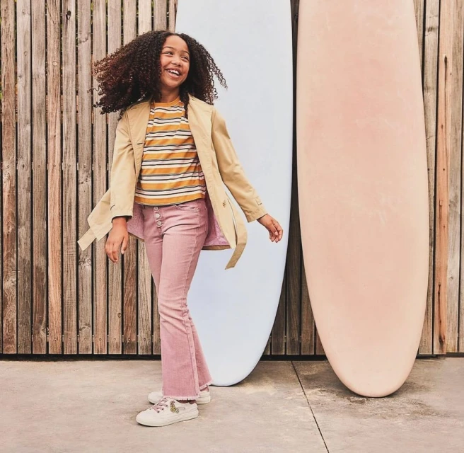 Smiling young girl with curly hair poses in front of surfboards wearing a tan jacket and pink pants, outdoor setting, full shot.