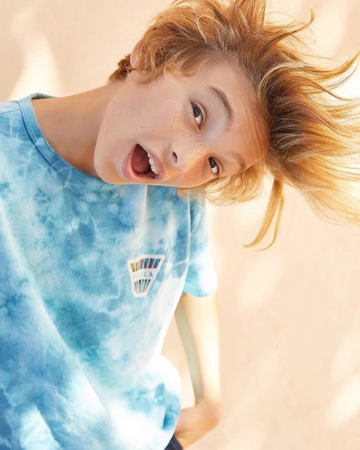 Young boy with blond hair wearing a tie-dye t-shirt with an open mouth, looking towards the camera, against a peach-colored backdrop.