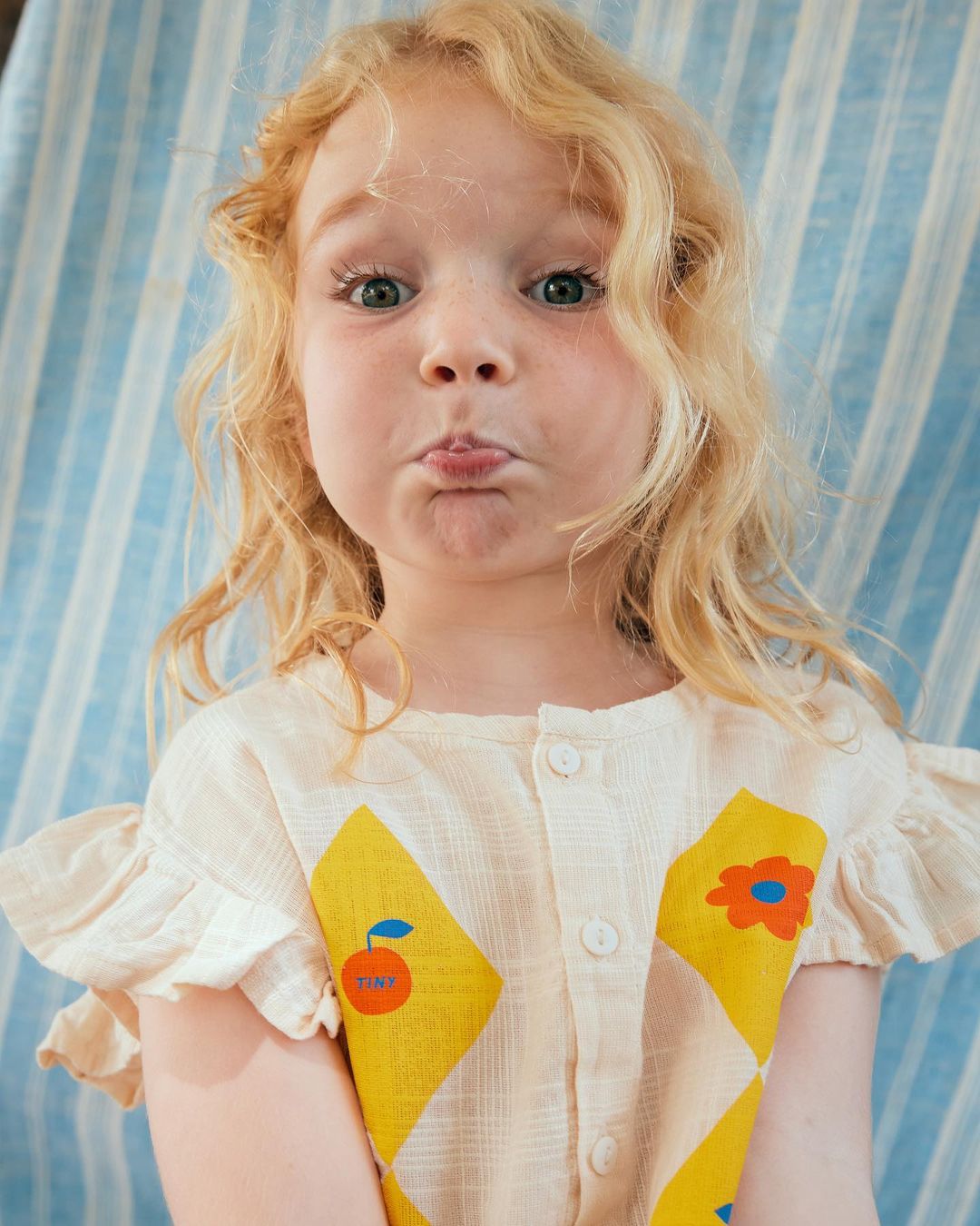 Portrait of a blonde child with a whimsical expression wearing a patterned romper against a vertically striped backdrop.