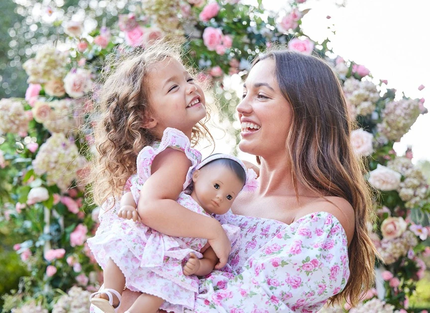 Woman and child smile in floral dresses, a doll, and a pink floral archway, creating a heartwarming scene in an outdoor setting.