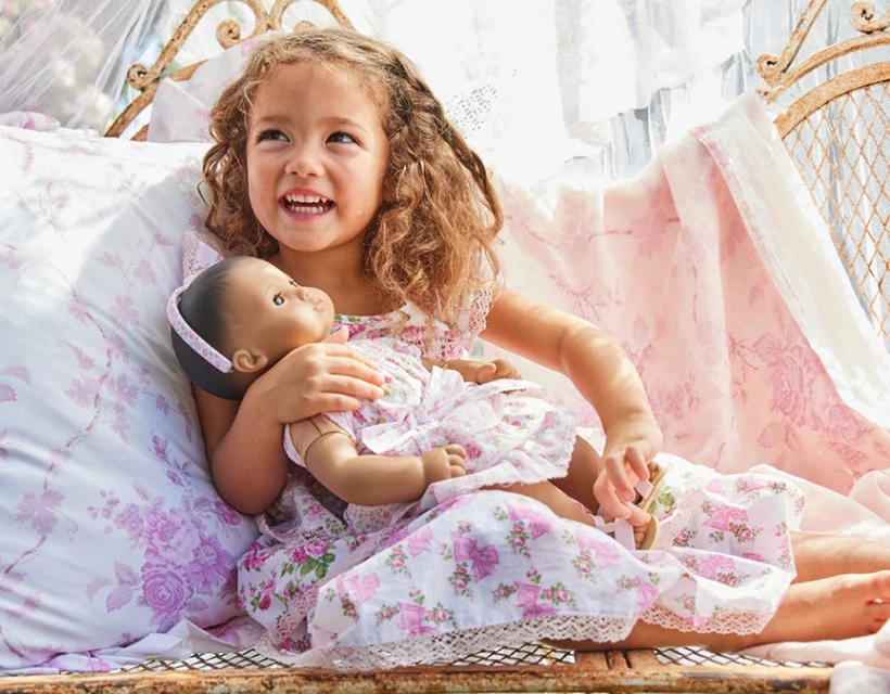 A young child with curly brown hair, wearing a floral dress, sits and smiles while holding a doll in a similar dress.
