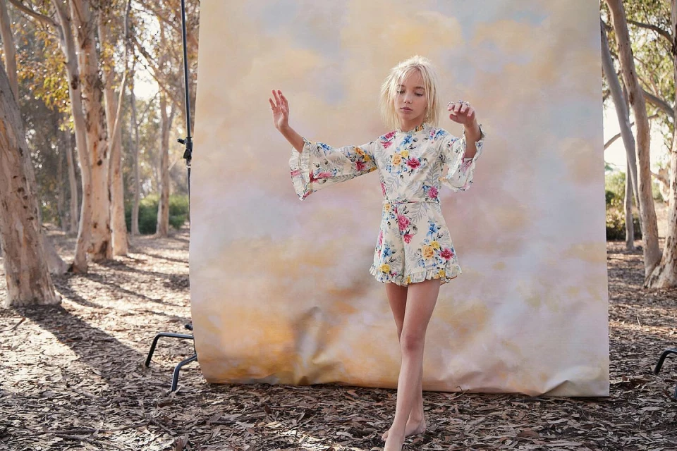 Young blonde girl poses in a floral romper outdoors against a painted backdrop, surrounded by trees in soft, natural light.