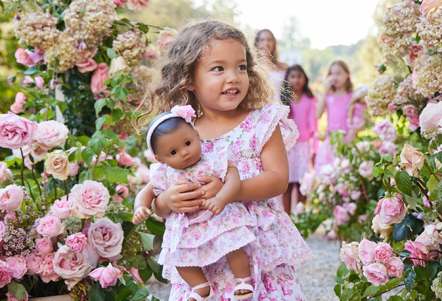 A young girl in a floral dress holds a doll in matching attire, surrounded by pink roses and other children in a garden setting.