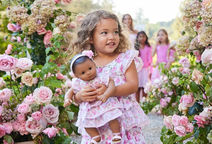 A young girl in a floral dress holds a doll in matching attire, surrounded by pink roses and other children in a garden setting.