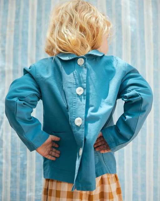 Child in blue jacket with white buttons, viewed from the back, wearing a plaid skirt against a striped blue and white backdrop.