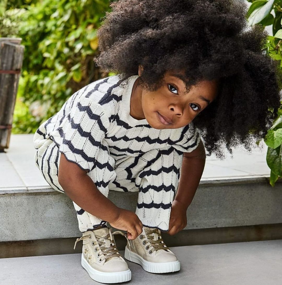 A young child with dark hair and fair skin kneels tying the laces of their neutral-colored high-top shoes outdoors with a green backdrop.