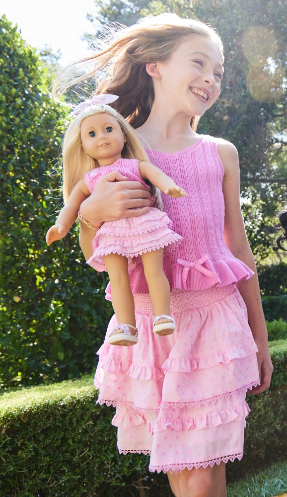Young girl with long hair and a pink dress smiles while holding a blonde-haired doll in a matching pink ruffled dress outdoors.