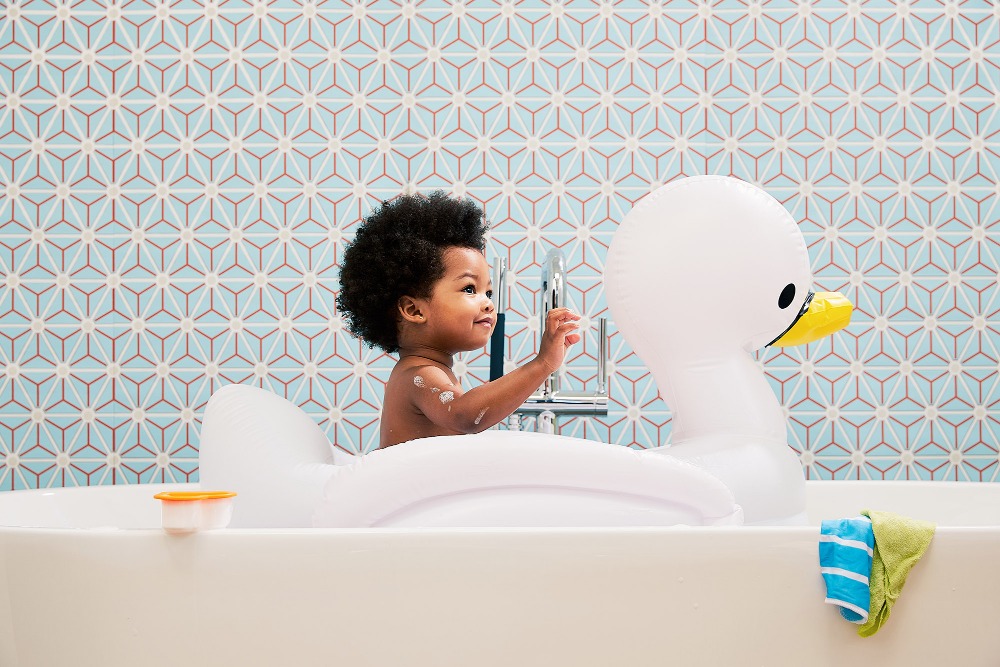 Toddler in bathtub with swan float and patterned tile backdrop, looking towards silver faucet.
