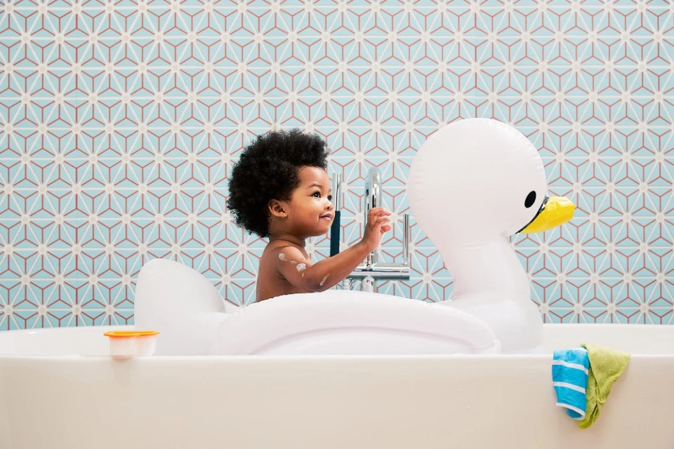 Toddler in bathtub with swan float and patterned tile backdrop, looking towards silver faucet.