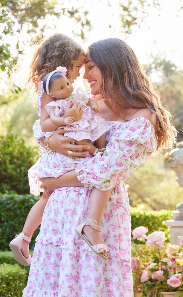 A happy mother in a floral dress holds her curly-haired daughter and a doll in a sunlit garden with flowers, creating a heartwarming scene.