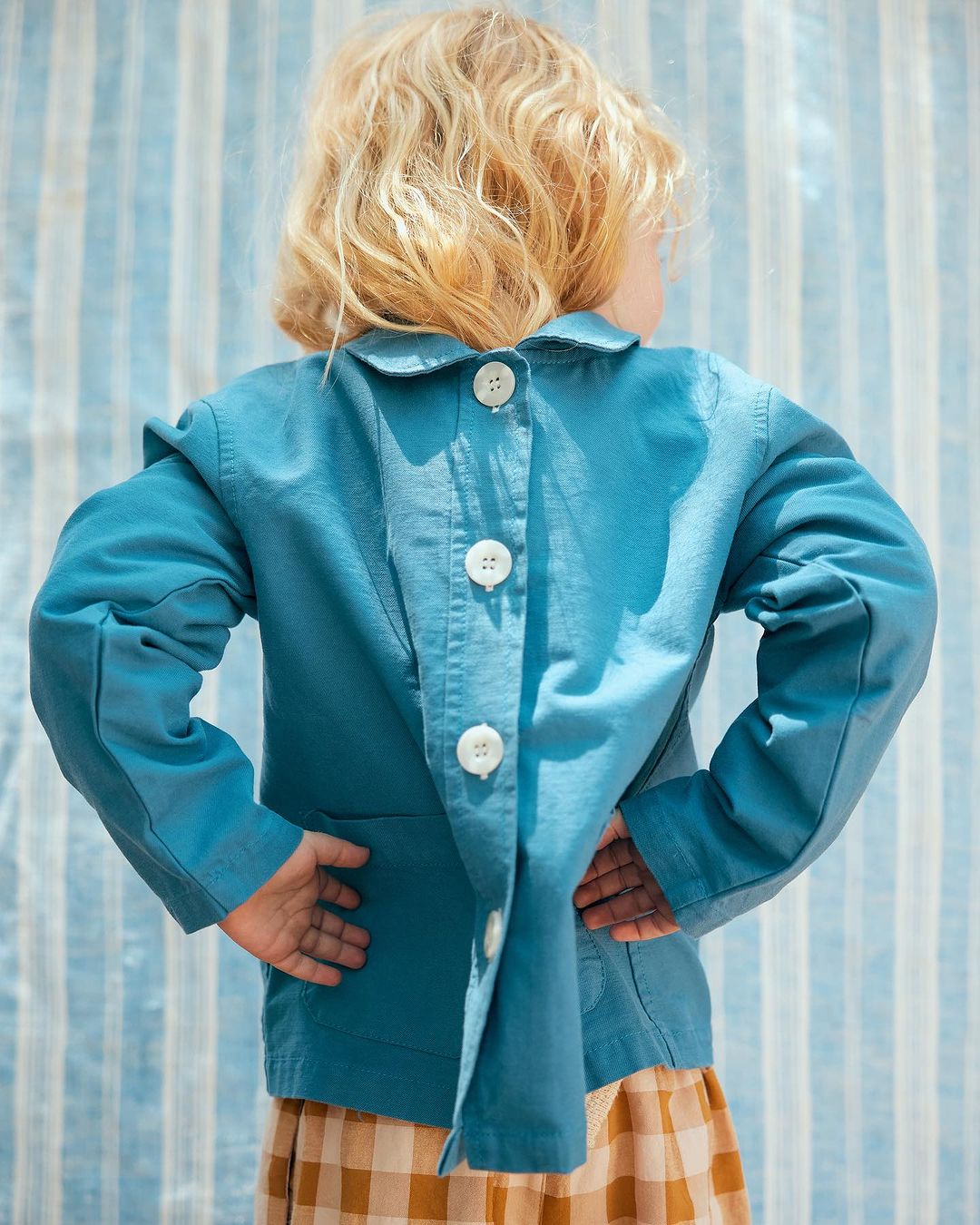 Back view of a young child wearing a teal collared jacket with white buttons and a tan and brown plaid skirt in a studio setting.