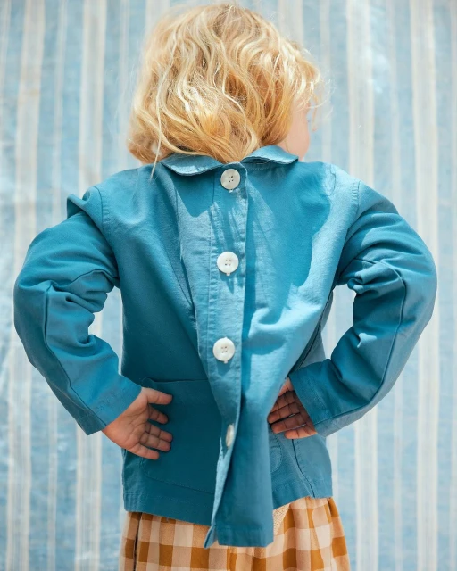 Back view of a young child wearing a teal collared jacket with white buttons and a tan and brown plaid skirt in a studio setting.