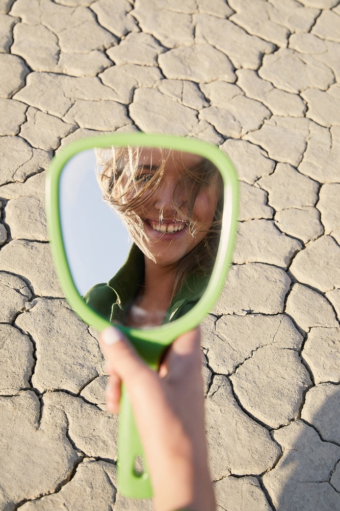 Woman's smiling face reflected in a handheld mirror against cracked desert ground. Holding a green mirror outside during the daytime.