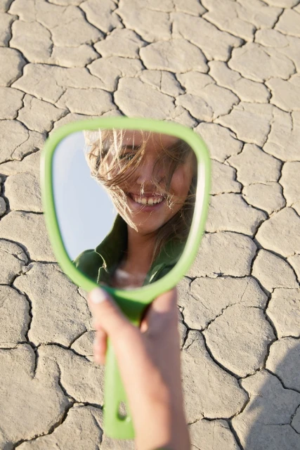 Woman's smiling face reflected in a handheld mirror against cracked desert ground. Holding a green mirror outside during the daytime.
