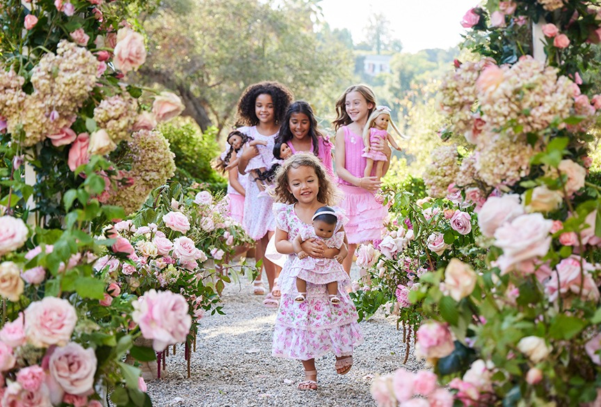 Four young girls in pink dresses walk through a flower-covered archway, each holding a doll, with greenery and a building in the background.