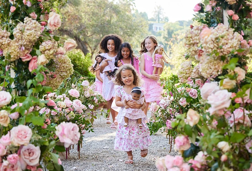 Four young girls in pink dresses walk through a flower-covered archway, each holding a doll, with greenery and a building in the background.