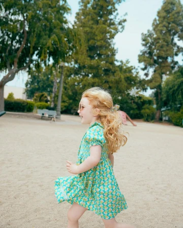 Young child with blonde curly hair wearing a patterned green dress running across a park path