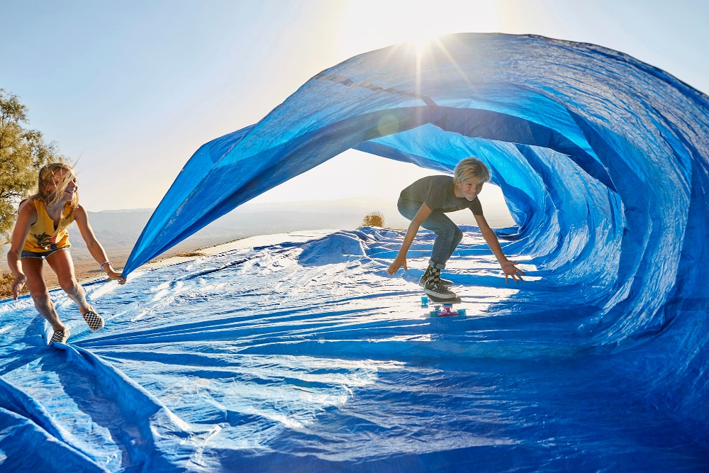 A youth on a skateboard is under a large blue tarp held by a woman on a sunny outdoor hilltop, creating a playful wave effect.