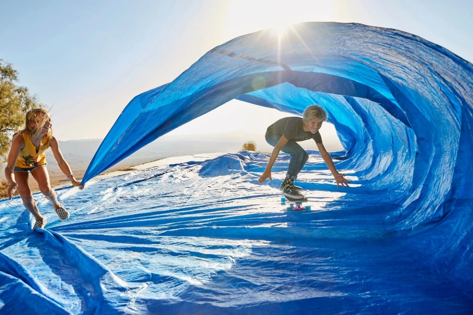 A youth on a skateboard is under a large blue tarp held by a woman on a sunny outdoor hilltop, creating a playful wave effect.
