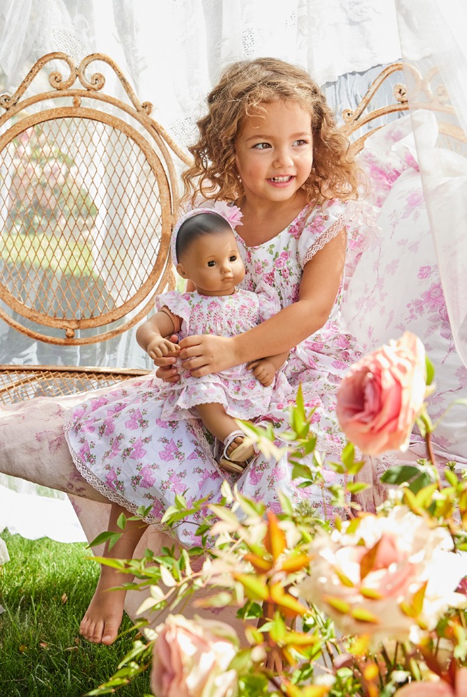A young girl with curly hair in a floral dress sits on an ornate metal daybed holding a doll surrounded by flowers in an outdoor setting.