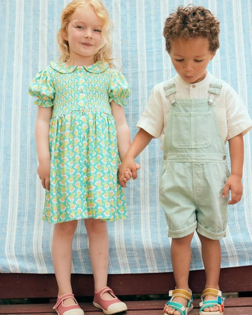 A young red-haired girl in a green floral dress and a young boy in green overalls hold hands while standing in front of a striped backdrop.