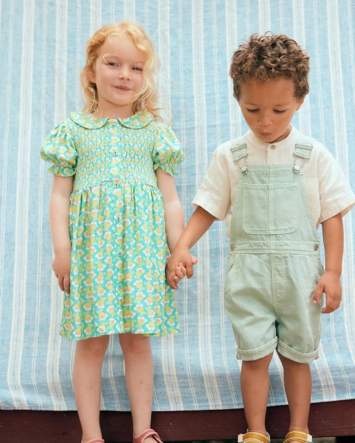 Two children, a girl and a boy, stand holding hands in front of a striped backdrop. The girl wears a patterned dress; the boy, overalls.