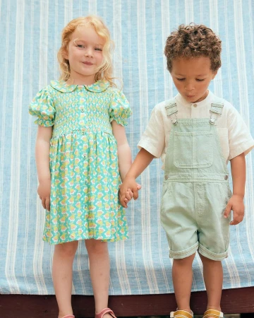Two children, a girl and a boy, stand holding hands in front of a striped backdrop. The girl wears a patterned dress; the boy, overalls.