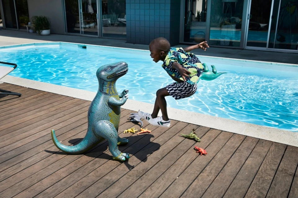 Child leaps near an inflatable dinosaur by a pool on a wood deck, captured in bright daylight.  Dinosaur toys scattered nearby.