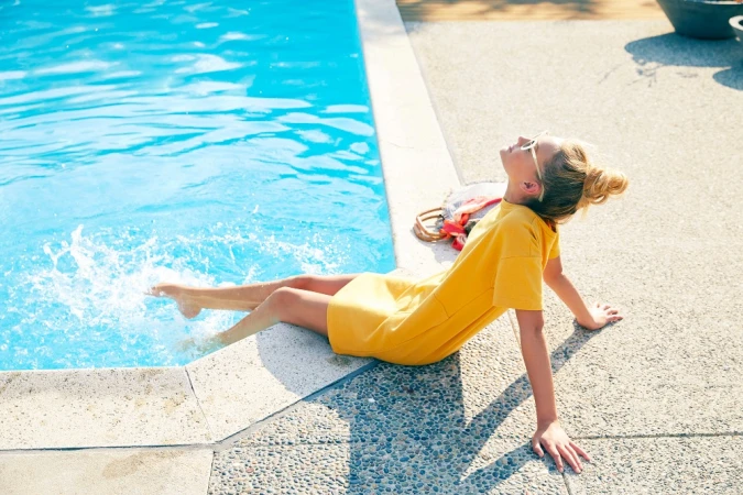 Young woman in a yellow dress sitting by a swimming pool, splashing water with her feet in Los Angeles by Don Diaz.