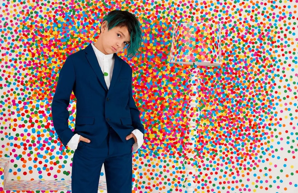Young boy in a blue suit poses in front of a colorful polka dot background with a matching lamp, creating a vibrant studio portrait.