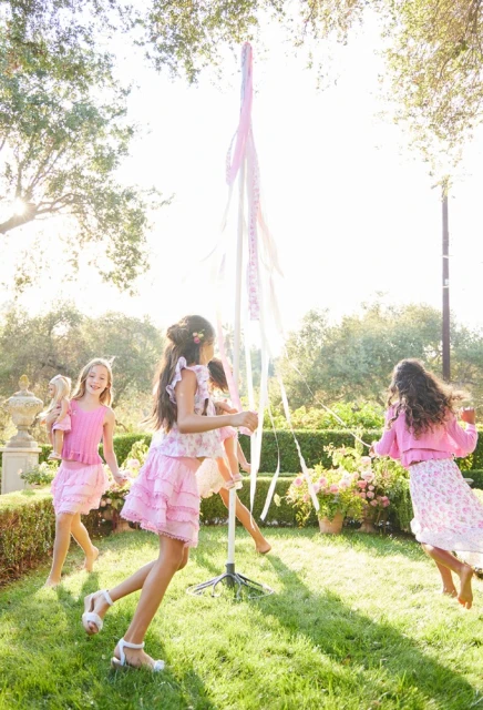 Four young girls play around a maypole in a grassy yard with flowering bushes and trees on a bright, sunny day outdoors.