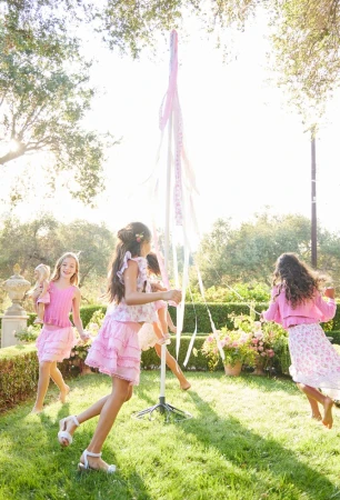 Four young girls play around a maypole in a grassy yard with flowering bushes and trees on a bright, sunny day outdoors.