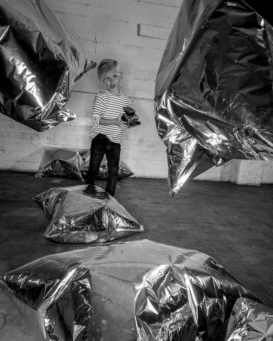 Black and white photo of a child in a striped shirt standing on a shiny inflated object and holding a camera indoors with bright lighting.