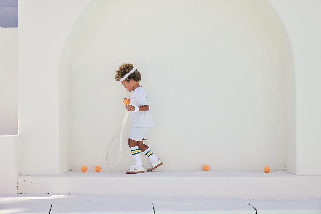 A child in tennis attire prepares to serve a tennis ball, standing on a white surface with tennis balls scattered nearby, bright sunny day.