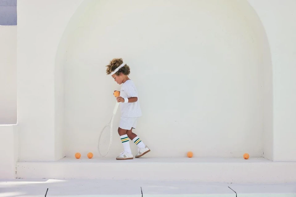 A child in tennis attire prepares to serve a tennis ball, standing on a white surface with tennis balls scattered nearby, bright sunny day.