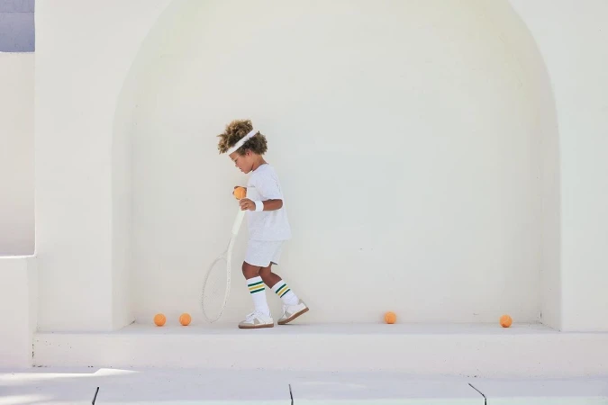 A child in tennis attire prepares to serve a tennis ball, standing on a white surface with tennis balls scattered nearby, bright sunny day.