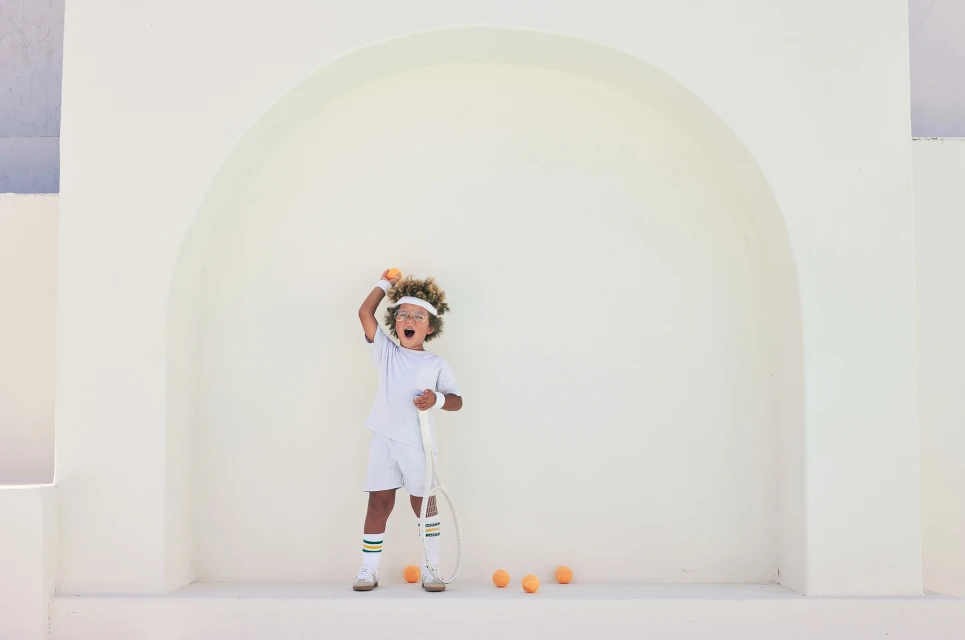 Child in vintage tennis outfit shouts in front of a white wall with tennis racket, wristband, headband, and scattered tennis balls.