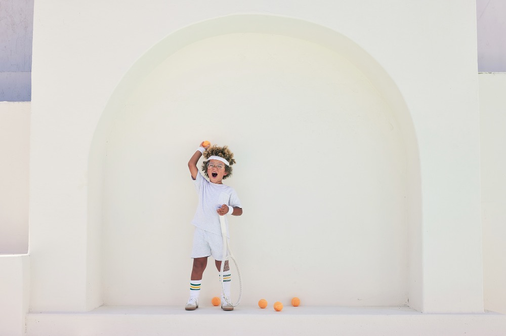 Young child in white tennis attire holding a tennis ball with a racket against a neutral arched background