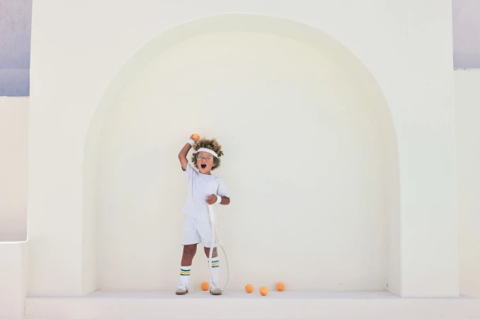 Young child in white tennis attire holding a tennis ball with a racket against a neutral arched background