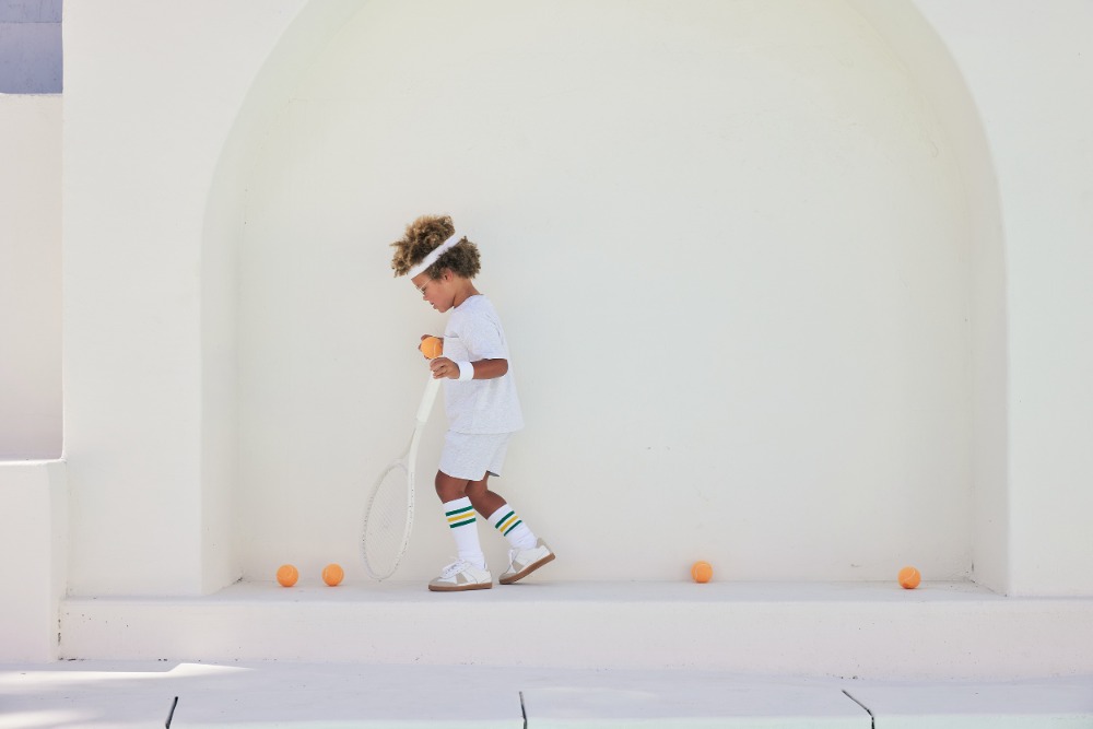 Young boy in white tennis outfit with racket and ball on a white architectural backdrop, surrounded by scattered tennis balls.