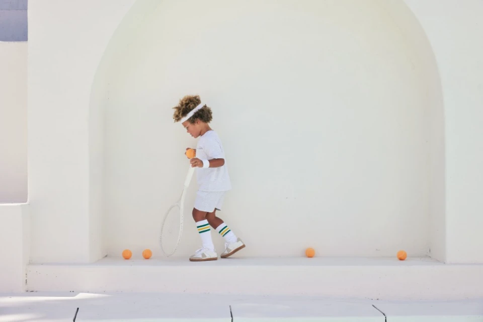 Young boy in white tennis outfit with racket and ball on a white architectural backdrop, surrounded by scattered tennis balls.