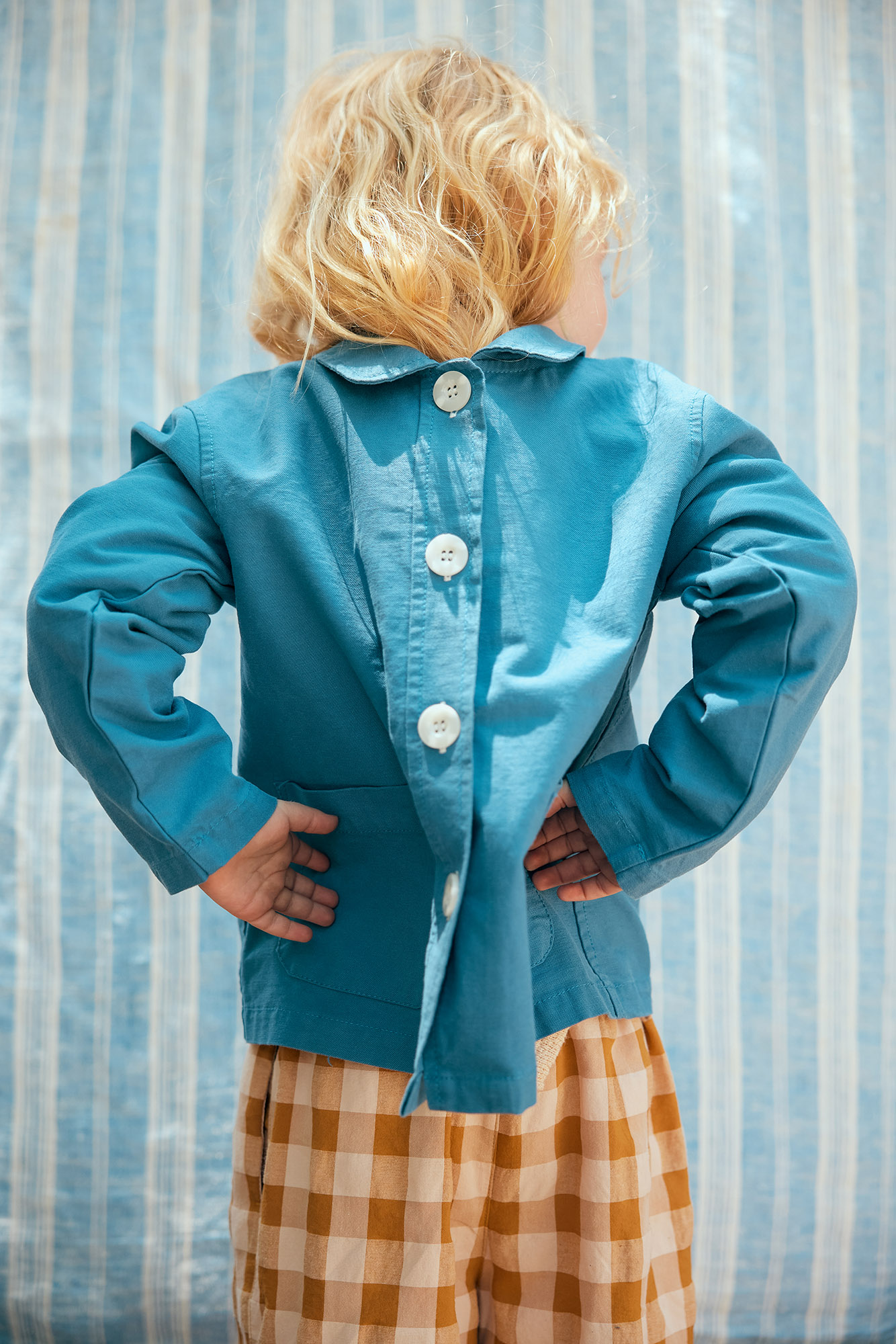 Back view of a toddler in a blue jacket and plaid pants, standing against a blue and white striped backdrop in a studio setting.