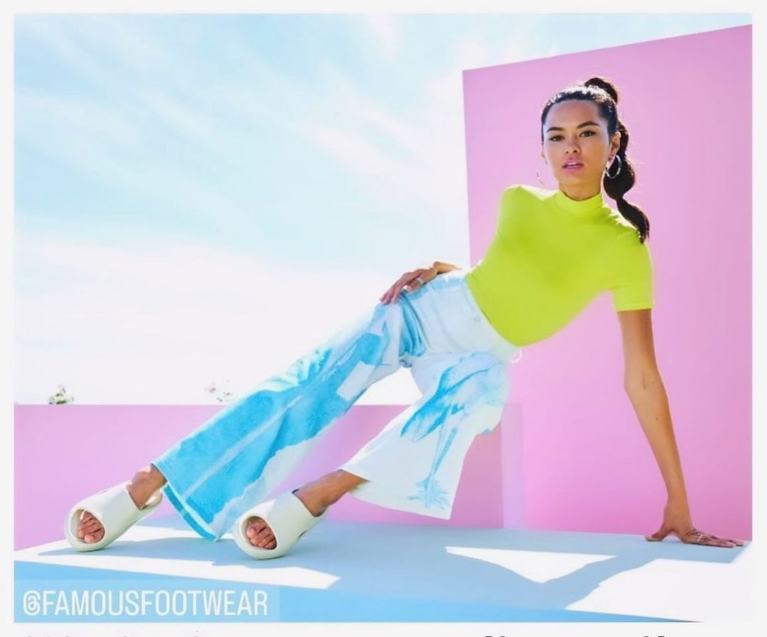 Woman poses in a neon yellow top and white pants with blue palm tree print, paired with platform slide sandals against a bright backdrop.